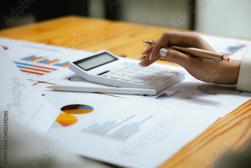 A businesswoman sits at her desk, calculating financial data on her laptop. Surrounded by paperwork and charts, she analyzes expenses and income, reflecting her role in corporate finance and accountin photo