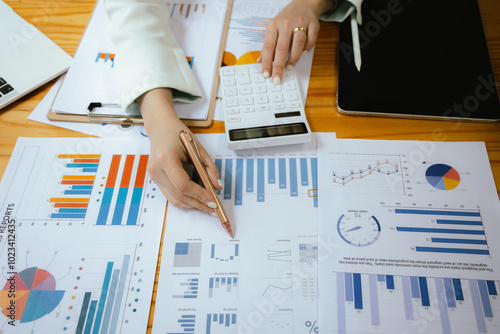 A businesswoman sits at her desk, calculating financial data on her laptop. Surrounded by paperwork and charts, she analyzes expenses and income, reflecting her role in corporate finance and accountin photo