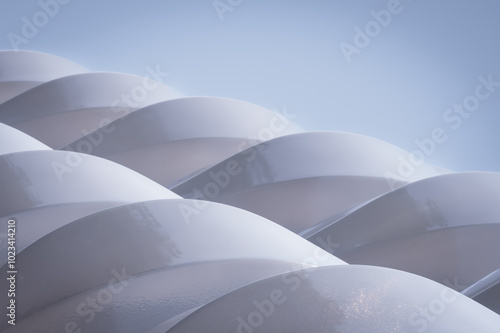 Curved, white structures are arranged in a repeating pattern against a soft blue background. Light reflects off the smooth surfaces, creating a serene and minimalist scene.