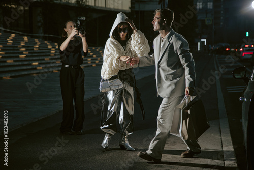 Fashionable man in grey classy suit helping girlfriend get out of car just arrived from shopping while paparazzi snapping photos of celebrity couple in city street at night, copy space