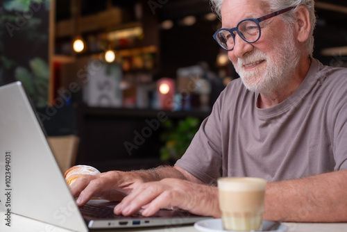 Handsome senior man enjoying breakfast with coffee and croissant sitting at a cafe while using laptop. Smiling bearded elderly male typing on keyboard from cafe in relaxed atmosphere