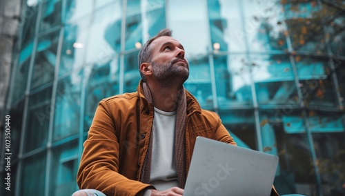 A contemplative man sits outside an urban building, using his laptop. The modern architecture reflects a blend of creativity and technology in city life.