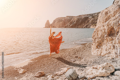 Woman red dress sea. Female dancer in a long red dress posing on a beach with rocks on sunny day. Girl on the nature on blue sky background.