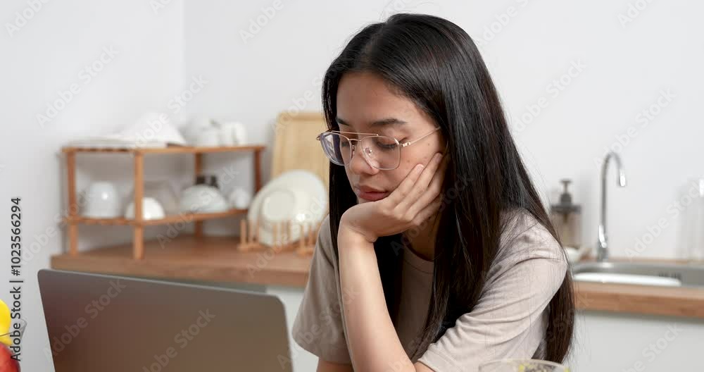 Asian woman resting chin on hand while looking at laptop in kitchen. Thoughtful expression, quiet atmosphere with glass of orange juice and clean kitchen background suggest work or study break. Asian woman resting chin on hand while looking at laptop in kitchen. Thoughtful expression, quiet atmosphere with glass of orange juice and clean kitchen background suggest work or study break.