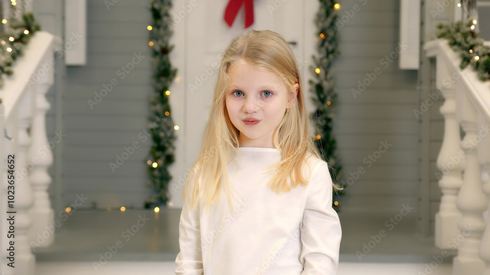 Fototapeta premium Blonde girl posing with a playful pout in a festive setting. A young blonde girl is seen playfully pouting while standing in front of a festive porch decorated with greenery and holiday lights.