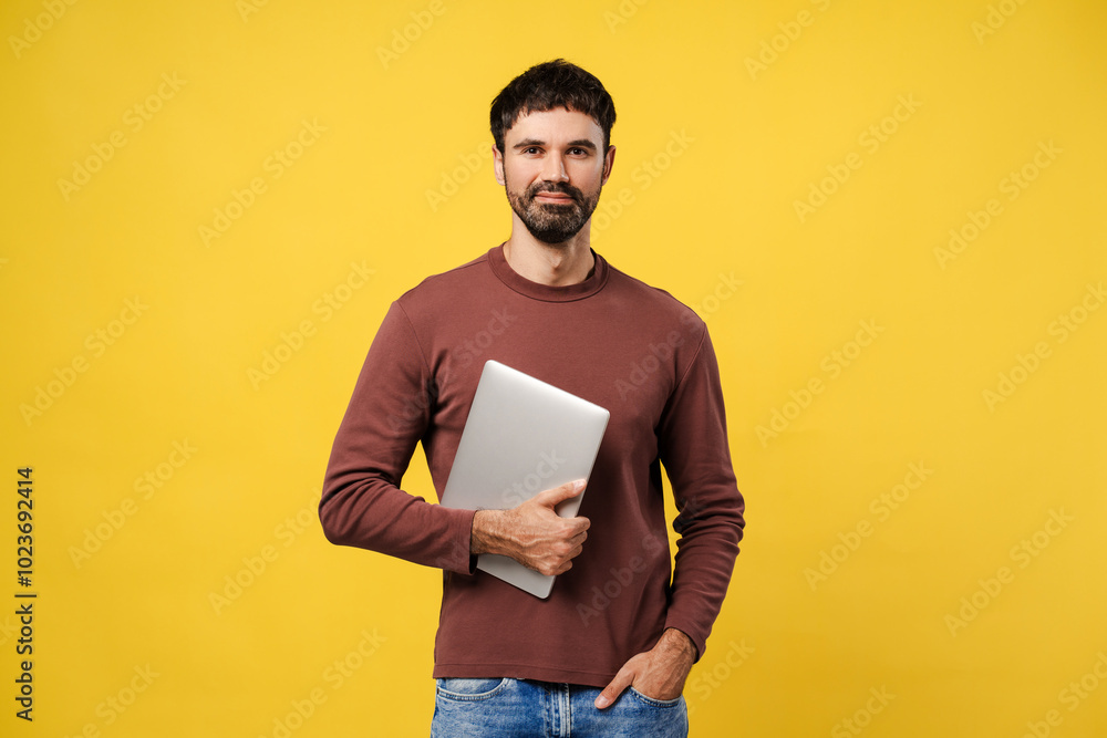 Fototapeta premium Success, confident man holding laptop, looking at camera standing isolated on yellow background