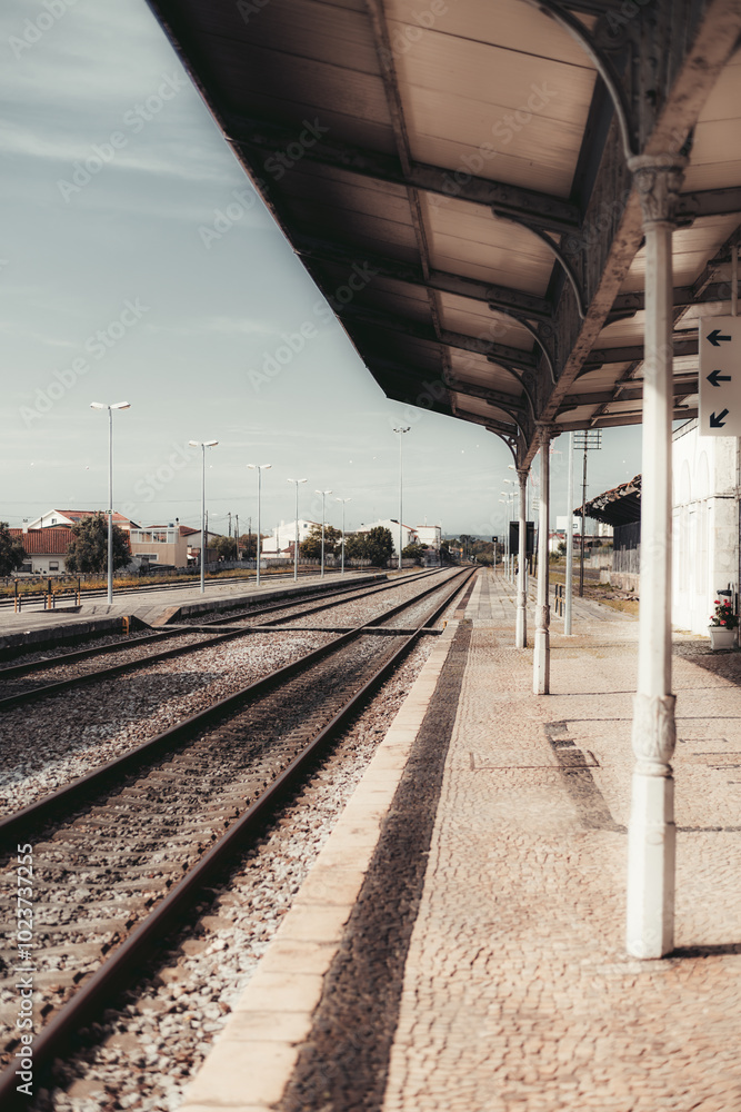 A vertical shot of a deserted train station platform with empty railway tracks extending into the distance. Covered shelter with metal support columns. Quiet urban surroundings under a clear sky A vertical shot of a deserted train station platform with empty railway tracks extending into the distance. Covered shelter with metal support columns. Quiet urban surroundings under a clear sky