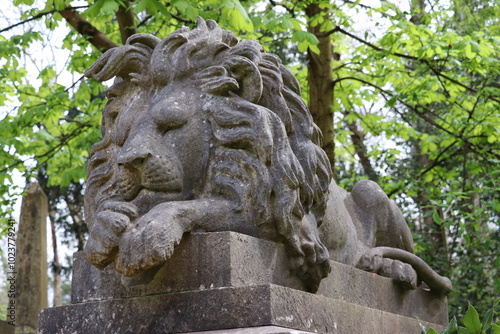 Photography A detailed stone lion sculpture rests on a pedestal amidst lush greenery in Highgate Cemetery, London, showcasing intricate carving and a serene expression