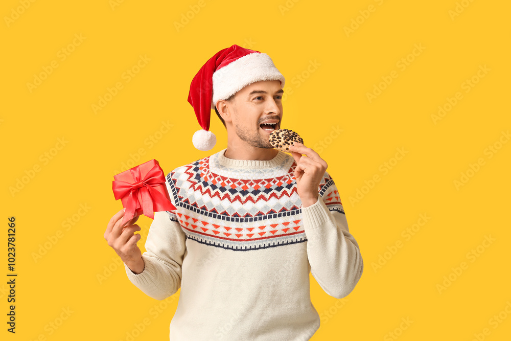 Handsome man in Santa hat with gift box and tasty cookie on yellow background Handsome man in Santa hat with gift box and tasty cookie on yellow background