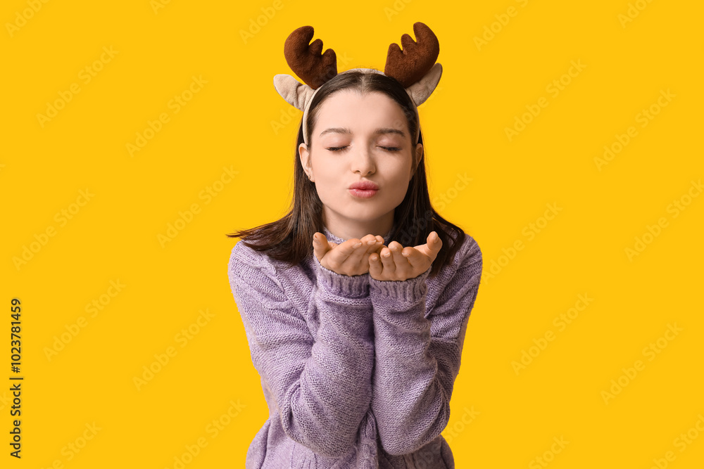 Beautiful young woman in reindeer horns blowing kiss on yellow background Beautiful young woman in reindeer horns blowing kiss on yellow background