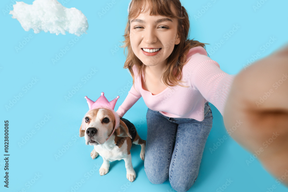 Young woman and cute beagle dog in princess crowns with white clouds taking selfie on blue background Young woman and cute beagle dog in princess crowns with white clouds taking selfie on blue background