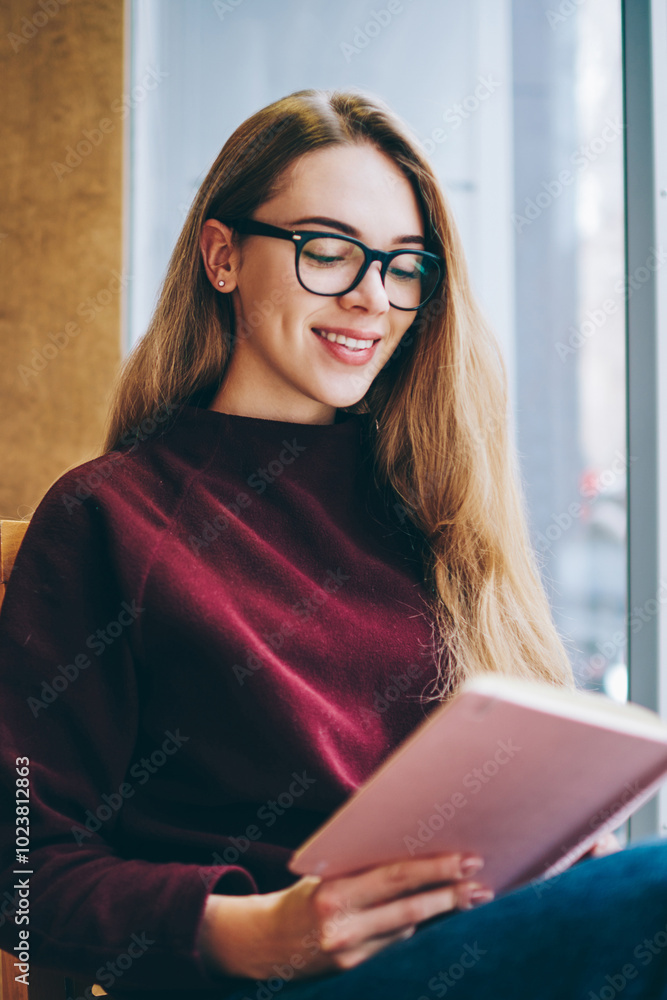 Cheerful female university student reading literature book in campus environment, positive teenager in spectacles for provide eyes protection sitting indoors and interested with open best seller Cheerful female university student reading literature book in campus environment, positive teenager in spectacles for provide eyes protection sitting indoors and interested with open best seller