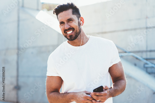 portrait of cheerful man dressed in blank t shirt enjoying free time for listening favourite music songs via electronic headphones,happy male with modern smartphone device smiling at camera photo