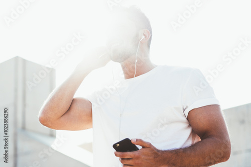 Below view of man with sunbeam on face enjoying music playlist weared in electronic headphones, millennial male meloman listening audio book via mobile knowledge application during leisure in city photo