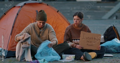 A homeless man and woman sit on the ground in front of a tent in the city. The man digs through his belongings while the woman holds a cardboard sign asking for help. Ideal for showcasing hardship.