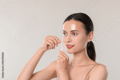 Smiling woman peeling off face mask on light grey background