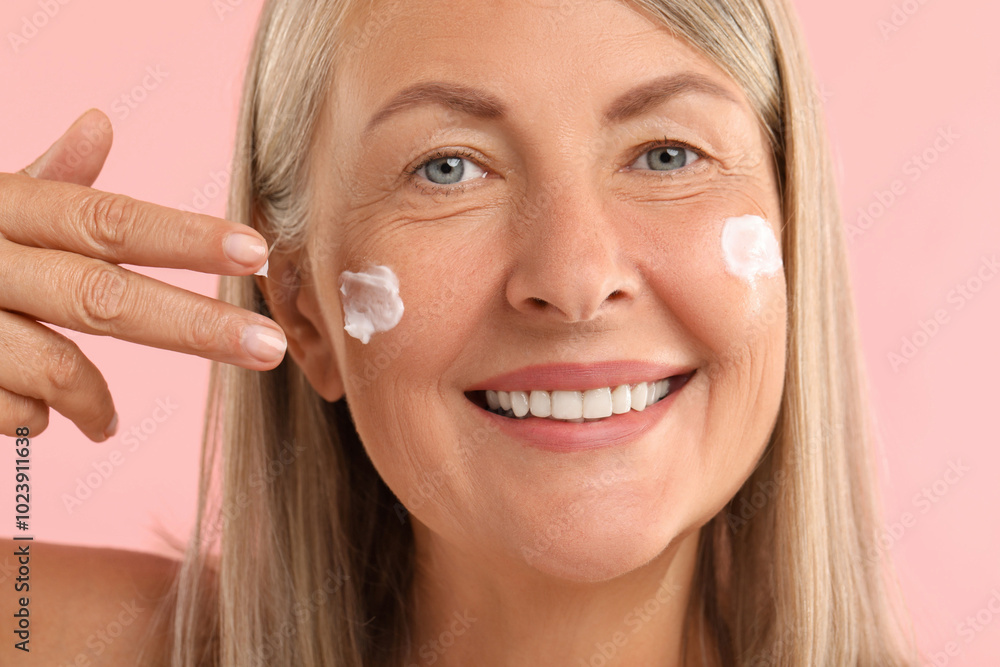Senior woman applying face cream on pink background, closeup Senior woman applying face cream on pink background, closeup