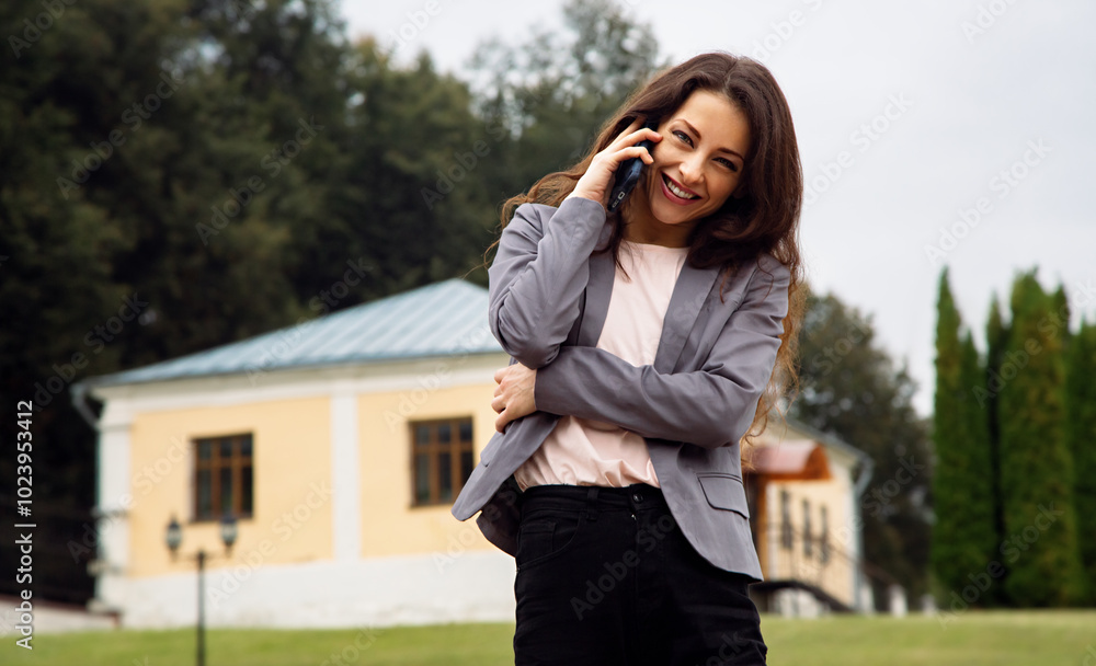 Fototapeta premium eautiful happy toothy smiling business woman in grey suit talking on the mobile phone walking outdoor nature summer green background