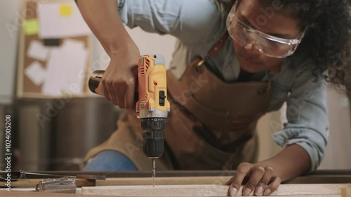Professional African American female carpenter worker wearing apron standing using electric power drill and screwdriver to assemble by screws to fix into the wooden at workplace in furniture factory. 