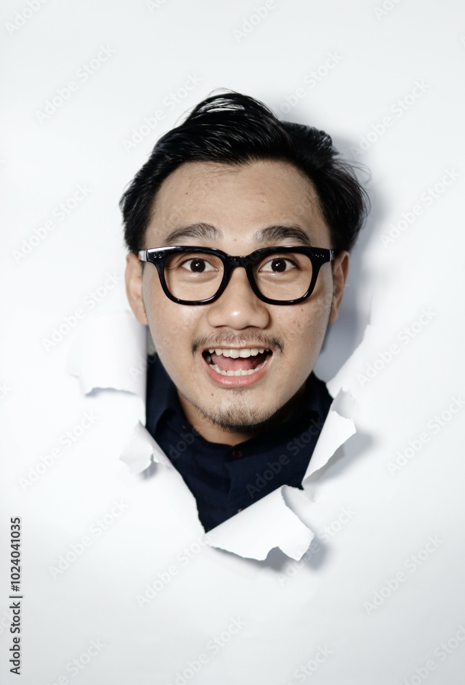 Happy asian man breaks through paper wall has fun looks gladfully at camera wears spectacles, smiles toothily enjoys making creative photos. Happy asian man breaks through paper wall has fun looks gladfully at camera wears spectacles, smiles toothily enjoys making creative photos.