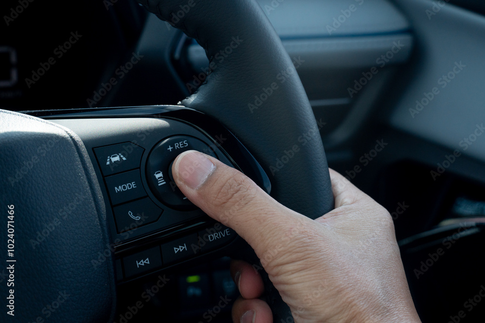 Hand of driver pressing a button on a car steering wheel. Steering wheel is black and has a leather or rubber grip. Hand is position in a way suggests the driver is using their thumb to press button. Hand of driver pressing a button on a car steering wheel. Steering wheel is black and has a leather or rubber grip. Hand is position in a way suggests the driver is using their thumb to press button.