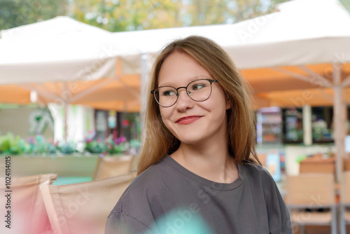 radiant, fair-haired teenage girl breaks into a warm smile as she sits at a table in a lively summer cafe, her joyful expression exuding a sense of relaxed contentment.