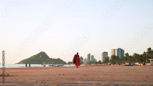 Hua Hin, Thailand. Lonely monk in orange kasaya clothes walking along a sandy beach at sunrise. The mountain and the city high-rise resort buildings in the background photo