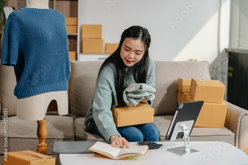 Asian lady blogger showing clothes in front of the camera to recording vlog video live streaming at her shop. Online Shopping Cart notion.