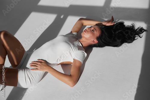Portrait of sporty athletic woman in white sportswear doing stretching, enjoying sport exercises, stretching legs, practicing yoga. Shot indoors with big window
