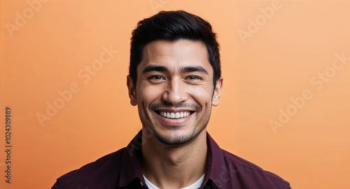 Young Latino man with narrow face grinning warm orange background