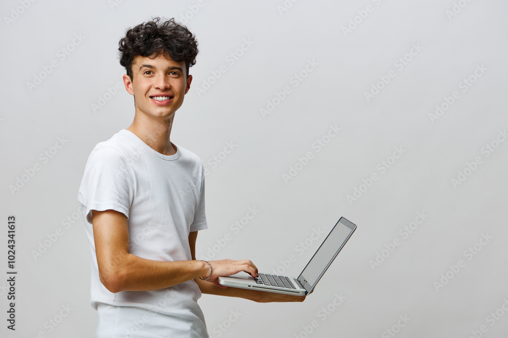 Young man smiling while using a laptop, casual attire, neutral background, positive technology concept, showcasing connection and creativity in personal or professional life Young man smiling while using a laptop, casual attire, neutral background, positive technology concept, showcasing connection and creativity in personal or professional life
