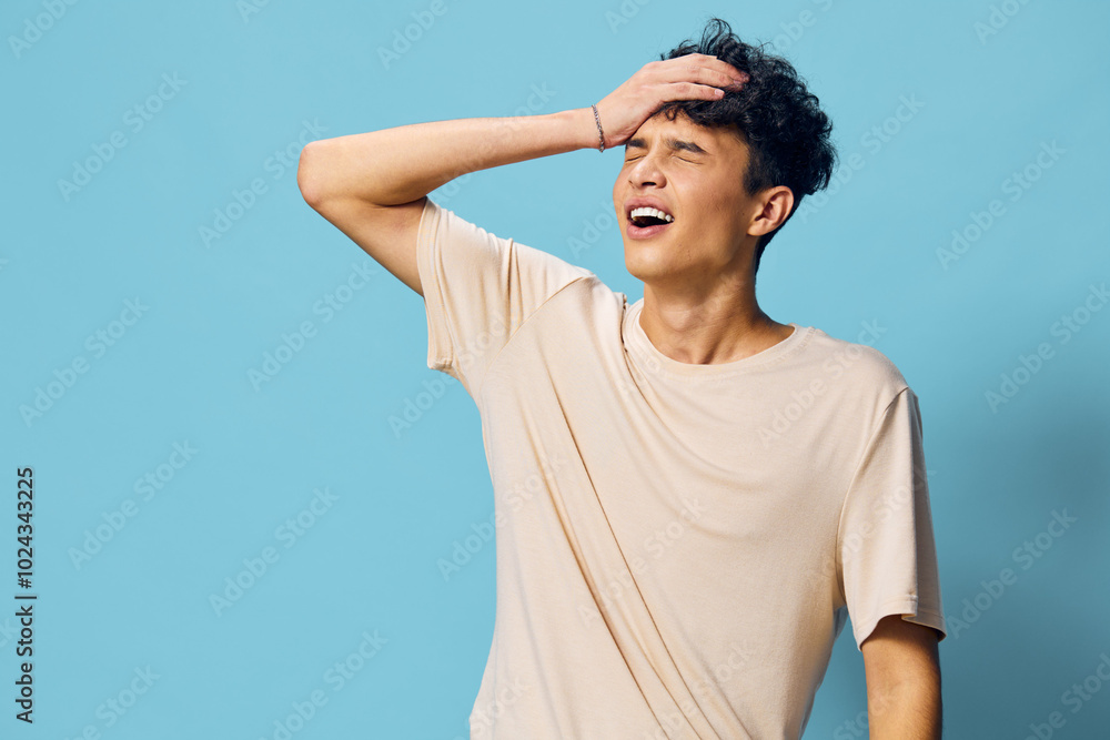 Young man with curly hair expressing frustration against a blue background, conveying emotions of stress and anxiety Casual beige shirt enhances the feeling of casual distress Young man with curly hair expressing frustration against a blue background, conveying emotions of stress and anxiety Casual beige shirt enhances the feeling of casual distress