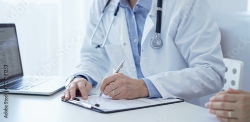 A doctor and a patient. The physician, wearing a white medical coat over a blue shirt, is filling out a medical record form during a consultation in the clinic. Medical service