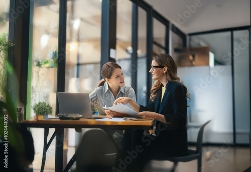 Two women are sitting at a table  discussing papers. One woman points to a document while the other listens attentively.