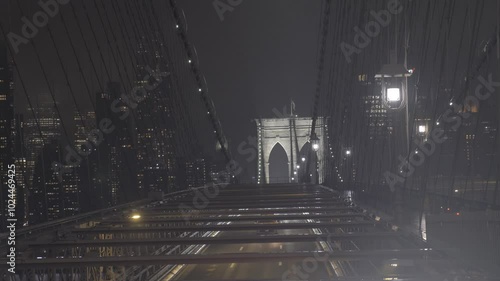 Cars passing by on the Brooklyn Bridge in New York, USA, filmed on a dark, cold, and foggy night, with the city's skyscrapers in the background.
