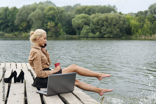 Obraz na płótnie Woman sitting on a pier by the river drinking coffee, talking on the phone and splashes water with his feet near the laptop w salonie