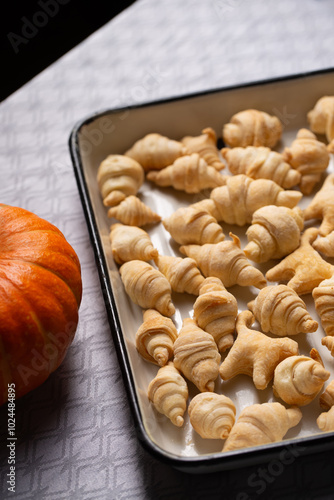 Small croissants and cookies in the shape of cats from puff pastry in a white baking dish, next to a pumpkin