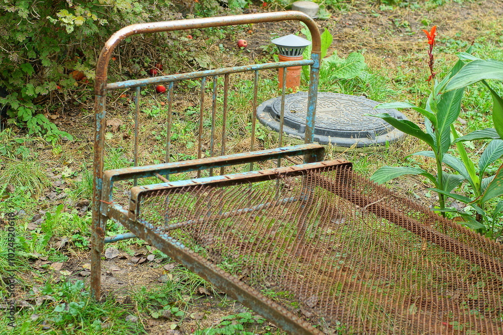 part of one painted old iron heavy blue industrial antique dirty rusty broken empty retro bed stands on the green grass near the leaves during the day on the street part of one painted old iron heavy blue industrial antique dirty rusty broken empty retro bed stands on the green grass near the leaves during the day on the street