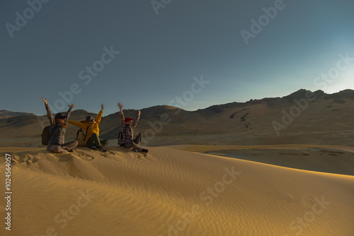 A group of people are sitting on the sand during the golden hour. Friends sit and raise their hands on the sand during sunset. A golden hour in the desert with people. Travelers are friends. Happy peo