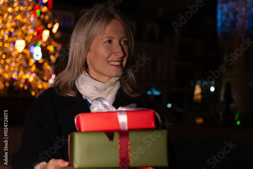 Young smiling blonde woman with gift boxes in her hands dreamily looks around against the background of a glowing Christmas tree at night on the street. Concept of Christmas, holidays