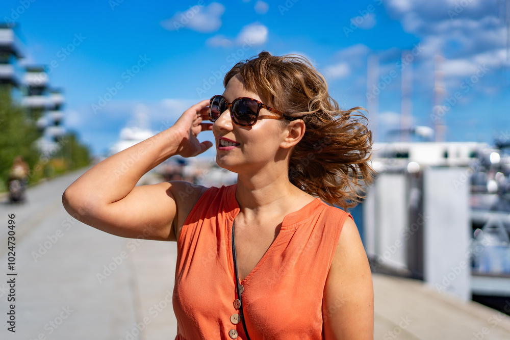 Sightseeing of seaside city. Portrait of middle-aged beautiful women wearing orange top and sunglasses looking out for something in yacht port in city on sunny day in summertime. Sightseeing of seaside city. Portrait of middle-aged beautiful women wearing orange top and sunglasses looking out for something in yacht port in city on sunny day in summertime.