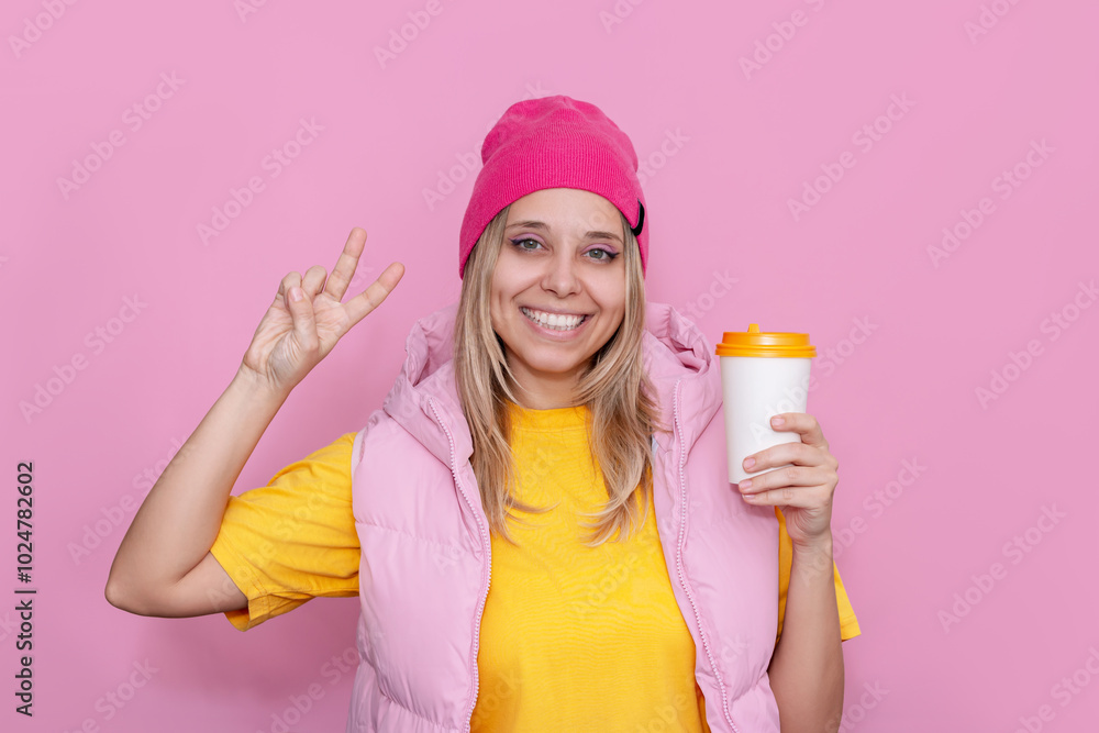 Young smiling blonde woman in a yellow T-shirt and bright pink hat holding a white paper cup of coffee or tea and showing peace gesture with her hand on a light pink color background Young smiling blonde woman in a yellow T-shirt and bright pink hat holding a white paper cup of coffee or tea and showing peace gesture with her hand on a light pink color background