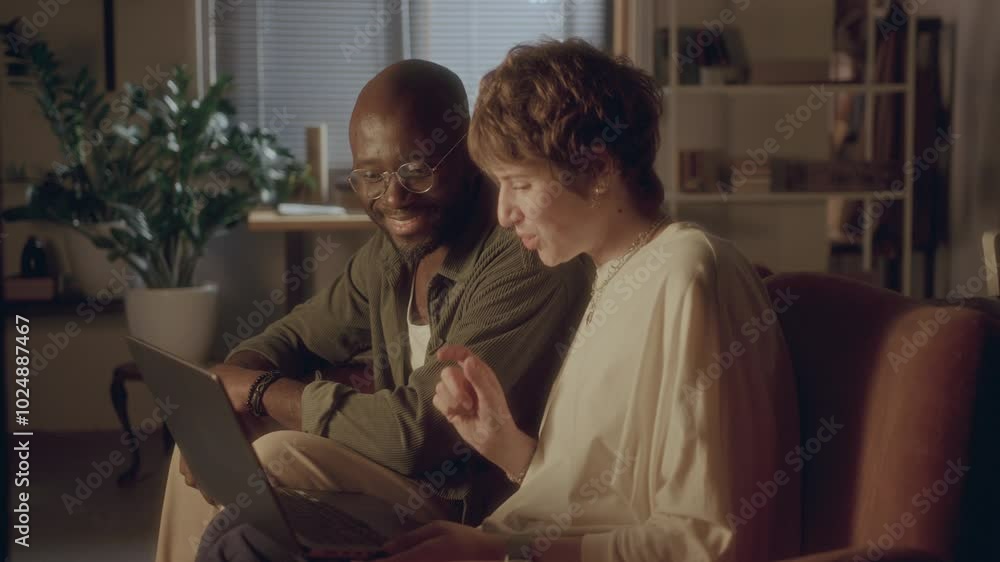 Black man and Caucasian woman sitting together on sofa in warmly lit living room, watching something on laptop screen, smiling and discussing it during evening at home Black man and Caucasian woman sitting together on sofa in warmly lit living room, watching something on laptop screen, smiling and discussing it during evening at home