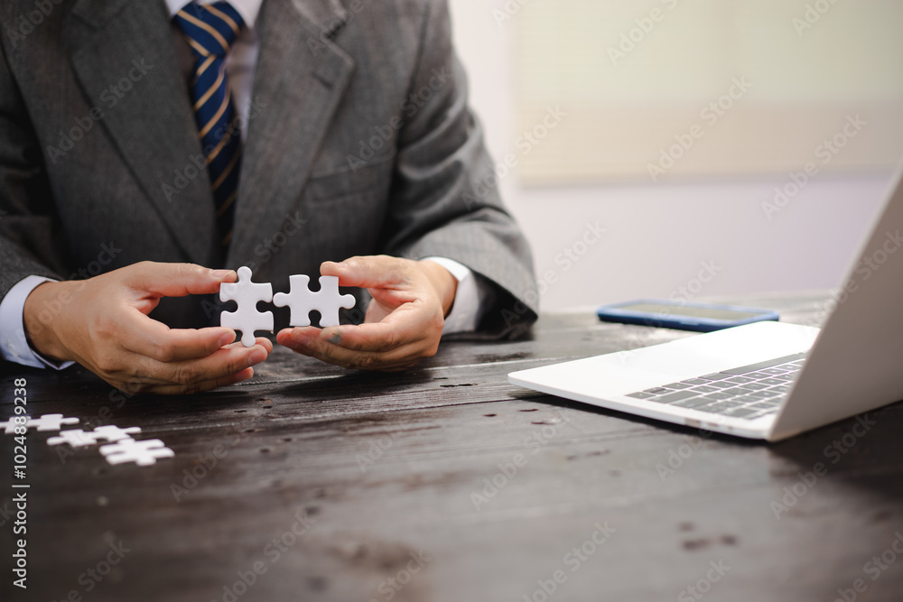 Businessman sitting at a table intently completing a jigsaw puzzle, demonstrating problem-solving, strategic thinking, and business decision-making, as well as attention to detail and patience. Businessman sitting at a table intently completing a jigsaw puzzle, demonstrating problem-solving, strategic thinking, and business decision-making, as well as attention to detail and patience.