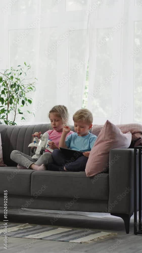Preschooler girl holds airplane toy while toddler boy looks in tablet. Siblings sit on sofa near big window watching educational lessons on gadget Preschooler girl holds airplane toy while toddler boy looks in tablet. Siblings sit on sofa near big window watching educational lessons on gadget