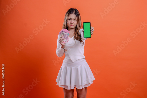 portrait of beautiful woman in white top standing annoyed to camera hand holding rupiah cash and cellphone screen, mobile phone in front isolated orange background. for service concept, business