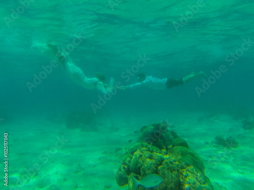 Beautiful young woman with long hair dressed in white swimsuit swims deep under the crystal azure water of caribbean sea, man diving underwater in black shorts