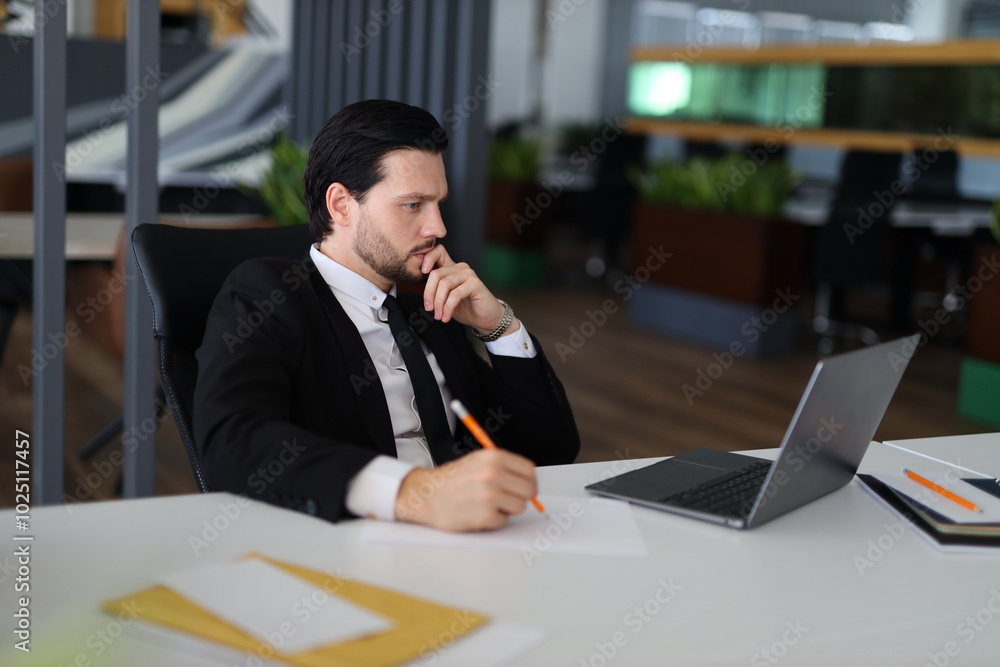 Fototapeta premium A man in a suit is sitting at a desk with a laptop and a pencil. He is writing something on a piece of paper. The scene suggests that he is working on a project or a report
