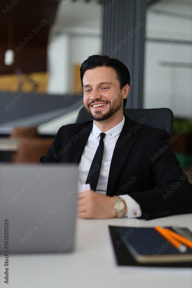 A man in a suit is sitting at a desk with a laptop and a book. He is smiling and he is enjoying his work A man in a suit is sitting at a desk with a laptop and a book. He is smiling and he is enjoying his work