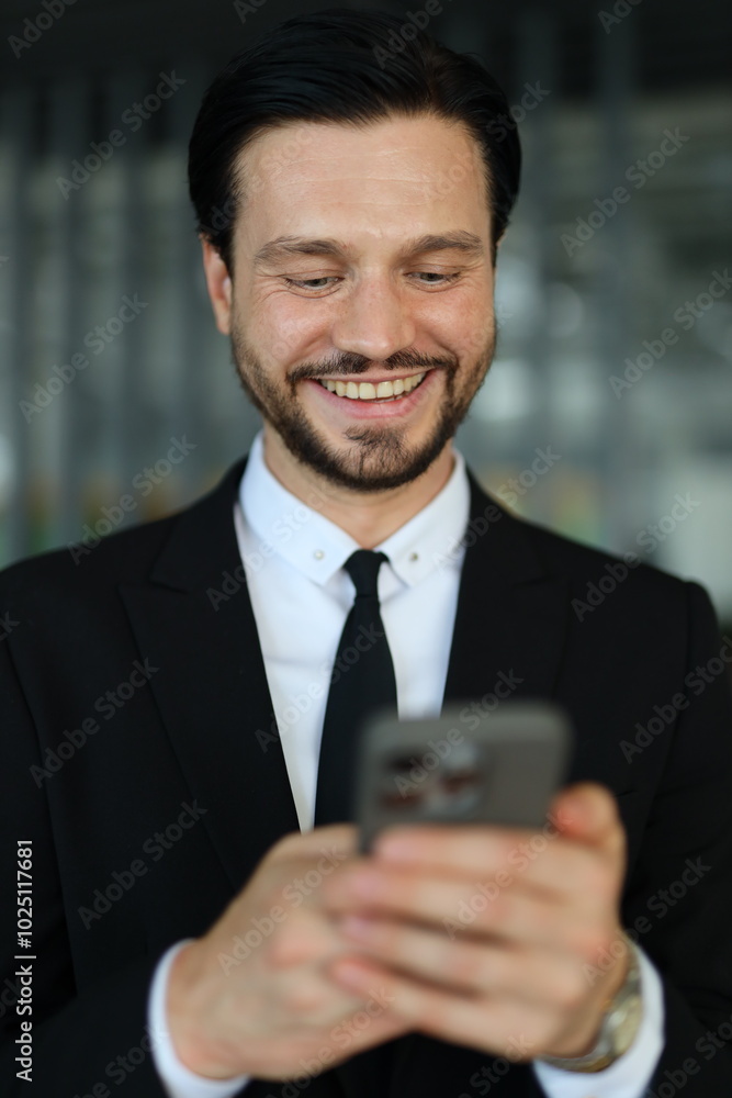 A man in a suit is smiling while looking at his cell phone. He is wearing a tie and he is in a professional setting A man in a suit is smiling while looking at his cell phone. He is wearing a tie and he is in a professional setting